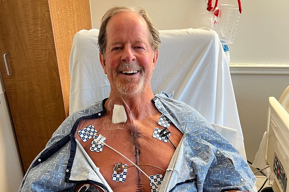 Cardiac surgery patient Eric Alcorn smiles in a hospital bed with a scar from his surgery visible between his gown