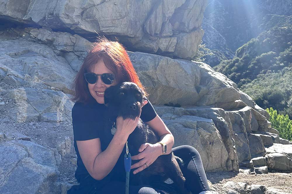 A redhaired woman cuddles a black dog in the mountains